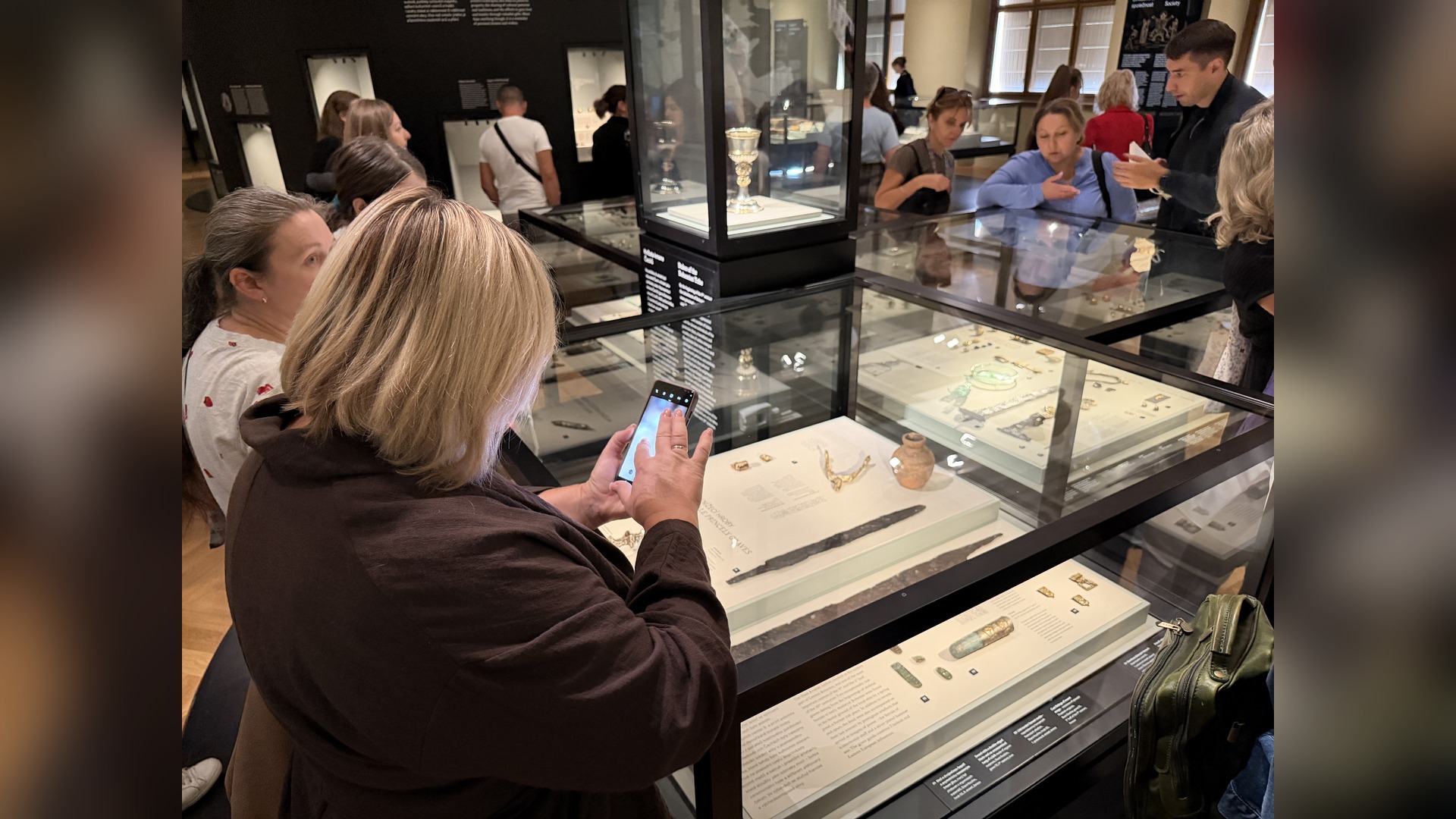 Program participants stand by glass display cases with exhibits, one of them photographing objects on her phone