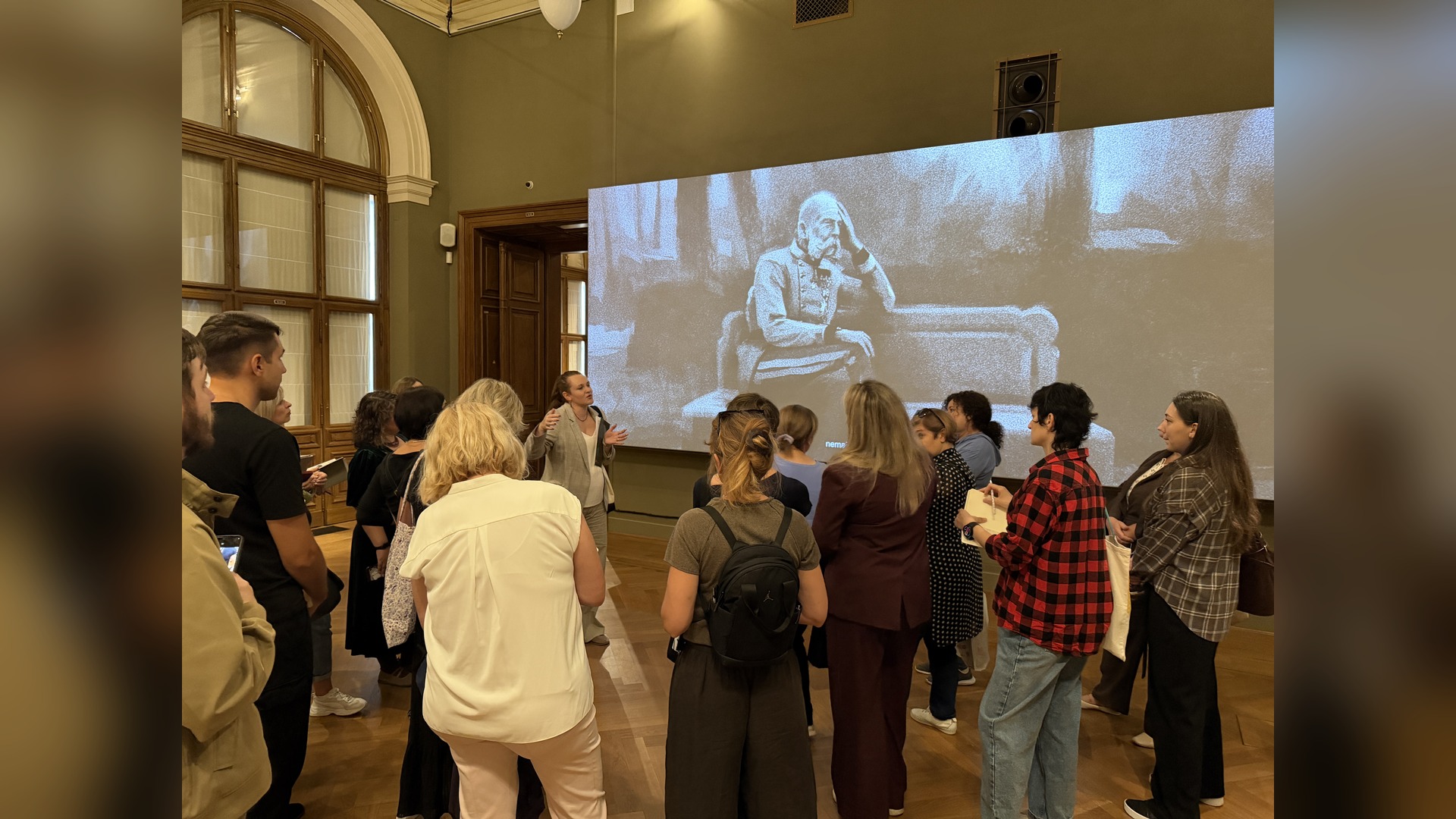 Program participants listen to a lecturer against the backdrop of a large historical black-and-white projection in the museum hall