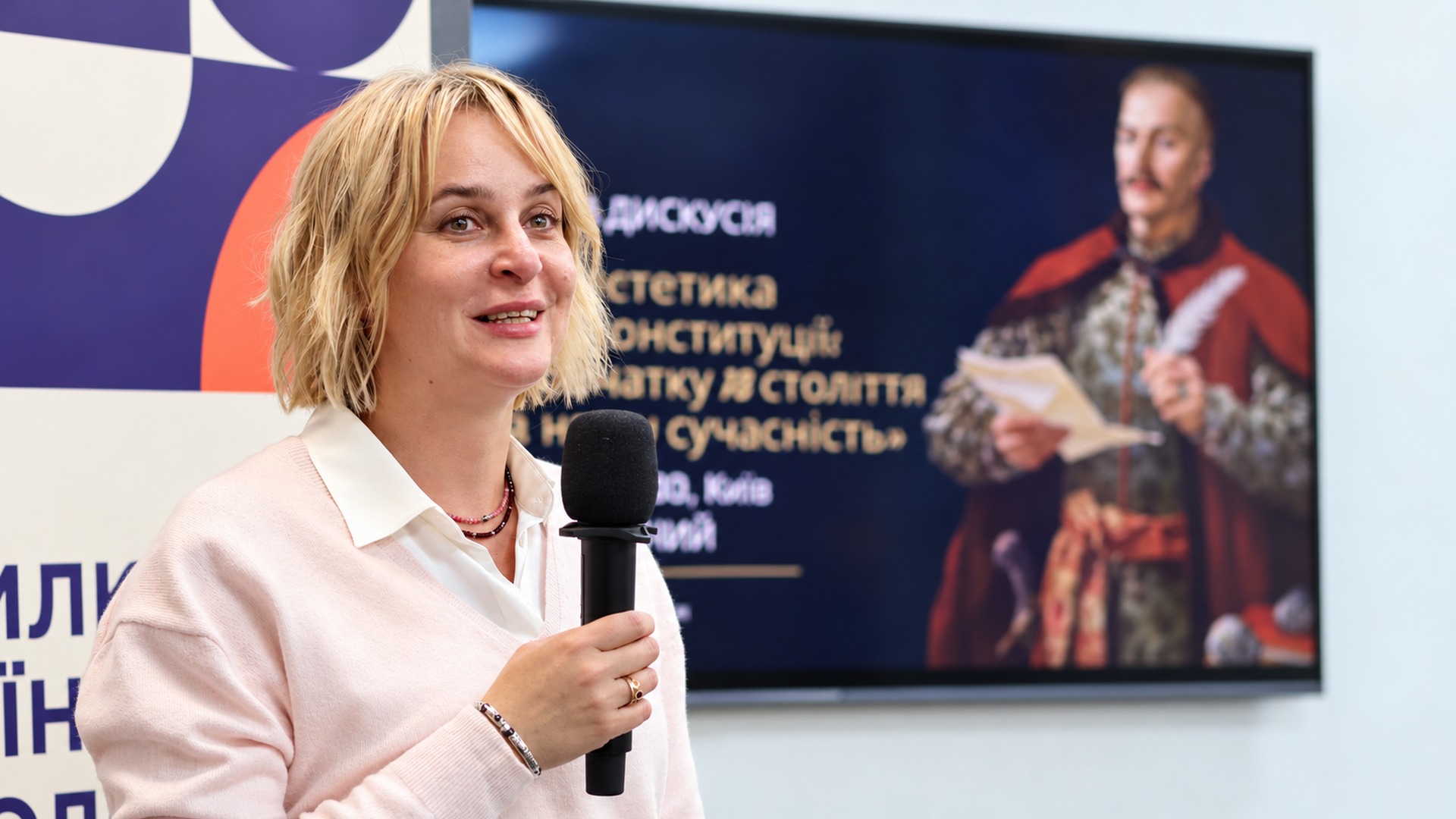 Svitlana Kvitnytska speaking into a microphone in front of a screen with a portrait of Hetman Pylyp Orlyk