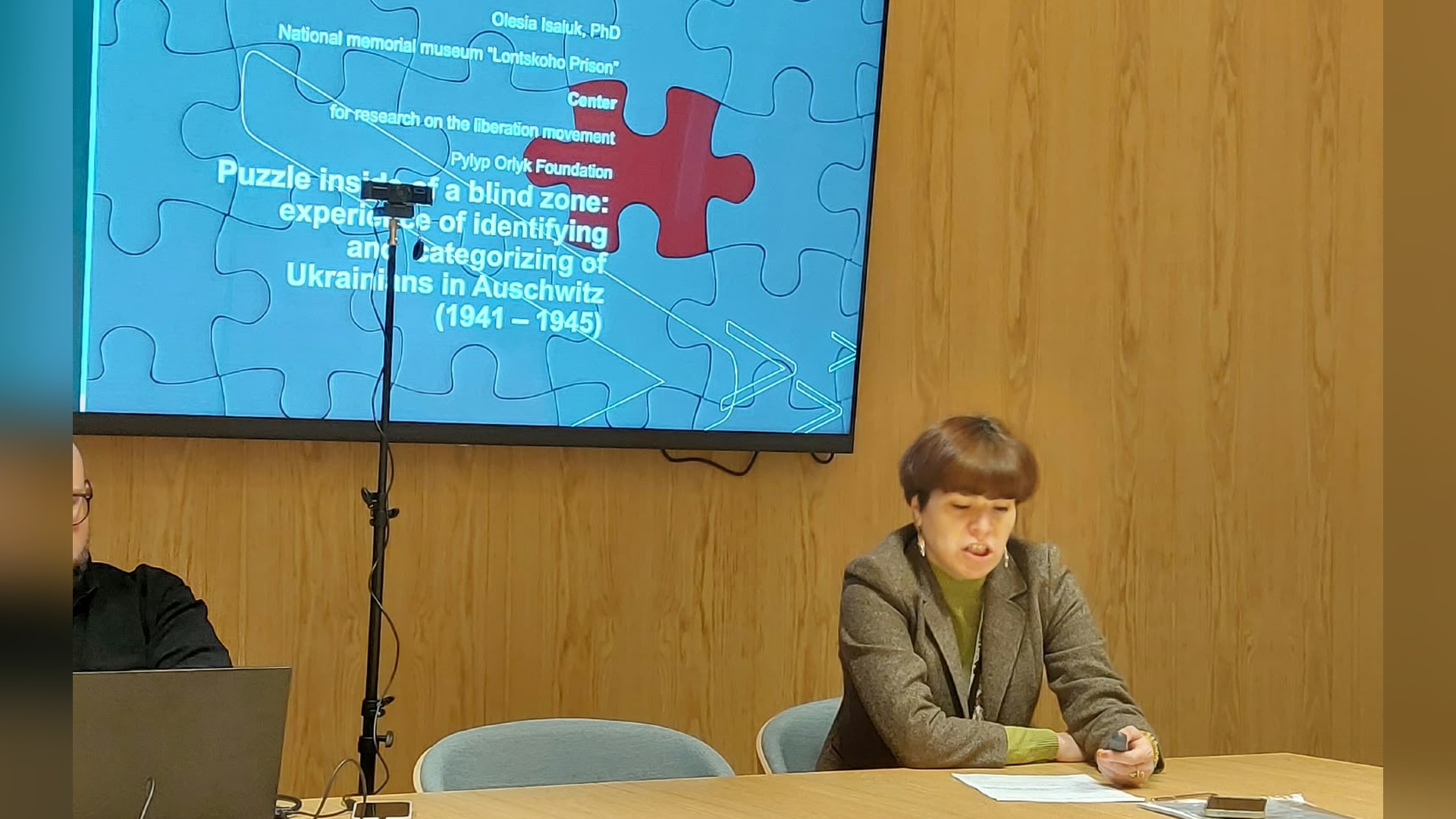 Олеся Ісаюк за столом представляє доповідь про ідентифікацію українців в Аушвіці, на екрані — слайд з пазлом