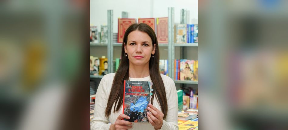 Ксенія Дворнікова тримає книгу «Вижити в Гостомелі: 21 історія з міста-героя» на тлі книжкових полиць