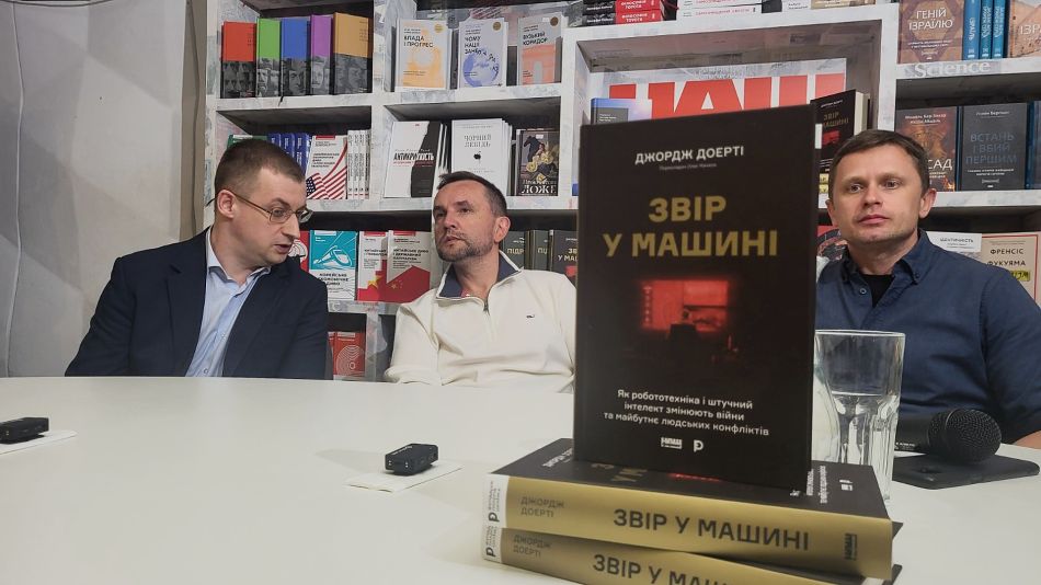 Троє учасників презентації за столом у книгарні, на передньому плані — книга Джорджа Доерті «Звір у машині»
