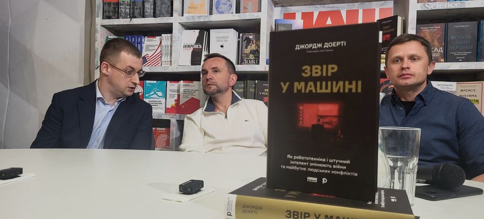 Троє учасників презентації за столом у книгарні, на передньому плані — книга Джорджа Доерті «Звір у машині»